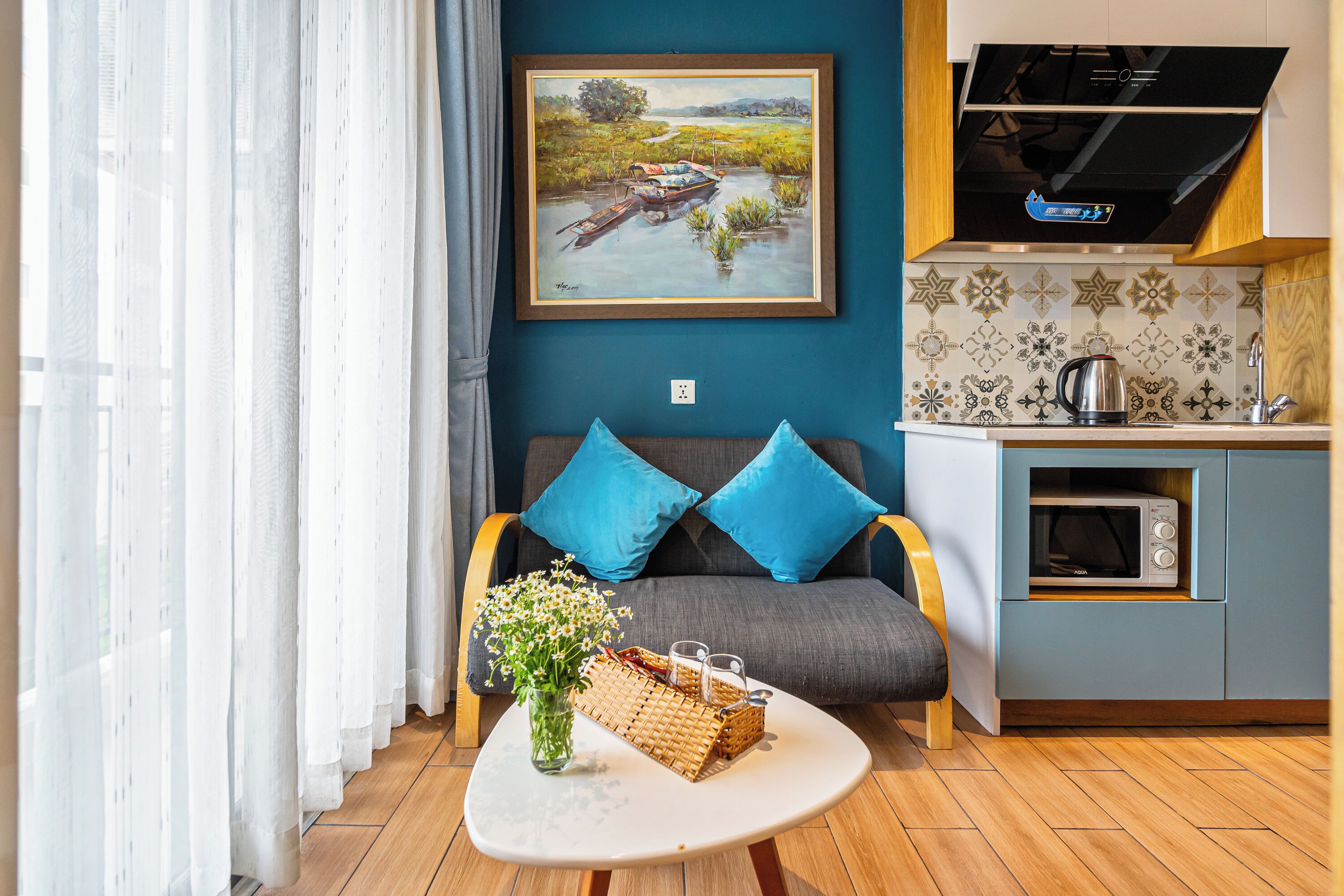 A modern living room with a sofa, a coffee table with flowers, and a kitchen area with a microwave and kettle.