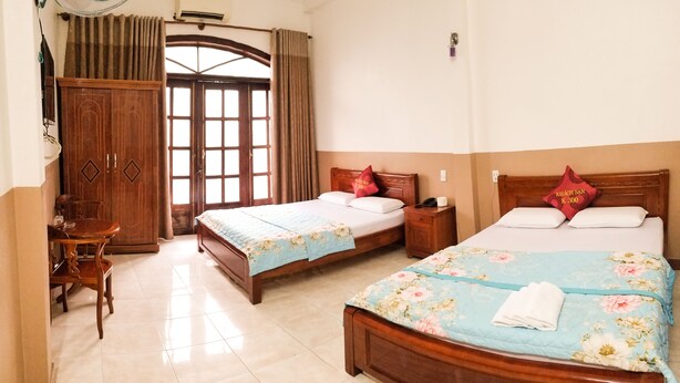 A hotel room with two beds, a wooden wardrobe, a desk with a chair, and a large window with curtains.