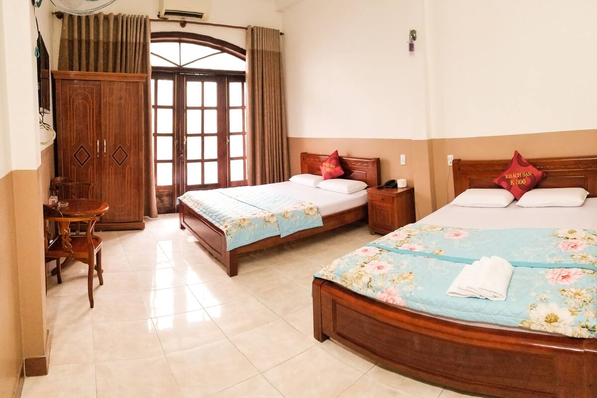 A hotel room with two beds, a wooden wardrobe, a desk with a chair, and a large window with curtains.
