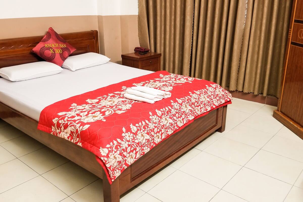 A neatly made bed with a red floral bedspread and white pillows, a wooden nightstand with a telephone, and a wooden wardrobe.