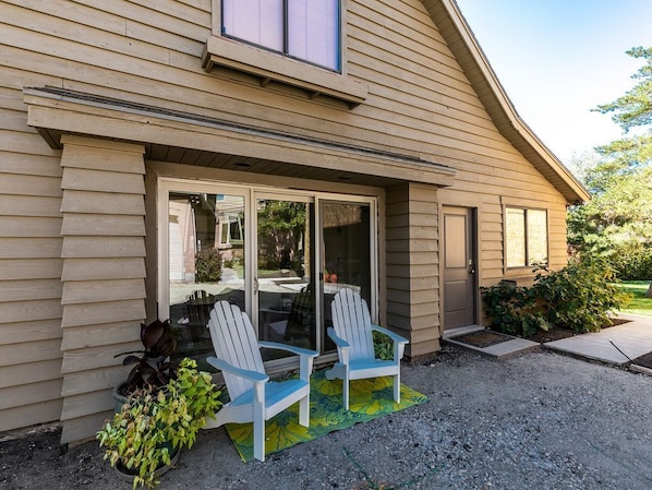 A house with a sliding glass door entrance, two Adirondack chairs outside, and a gravel driveway.
