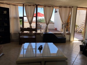 Une pièce comprenant une table à plateau en verre, un canapé, un fauteuil, une petite bibliothèque et donnant sur la terrasse extérieure.