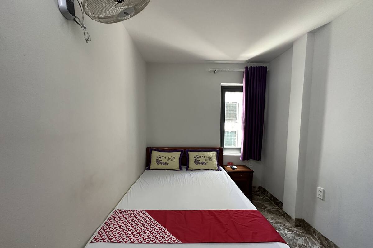 Ein schmales Hotelzimmer mit einem Bett, einem Nachttisch, einem Wandventilator und einem Fenster mit violetten Vorhängen.