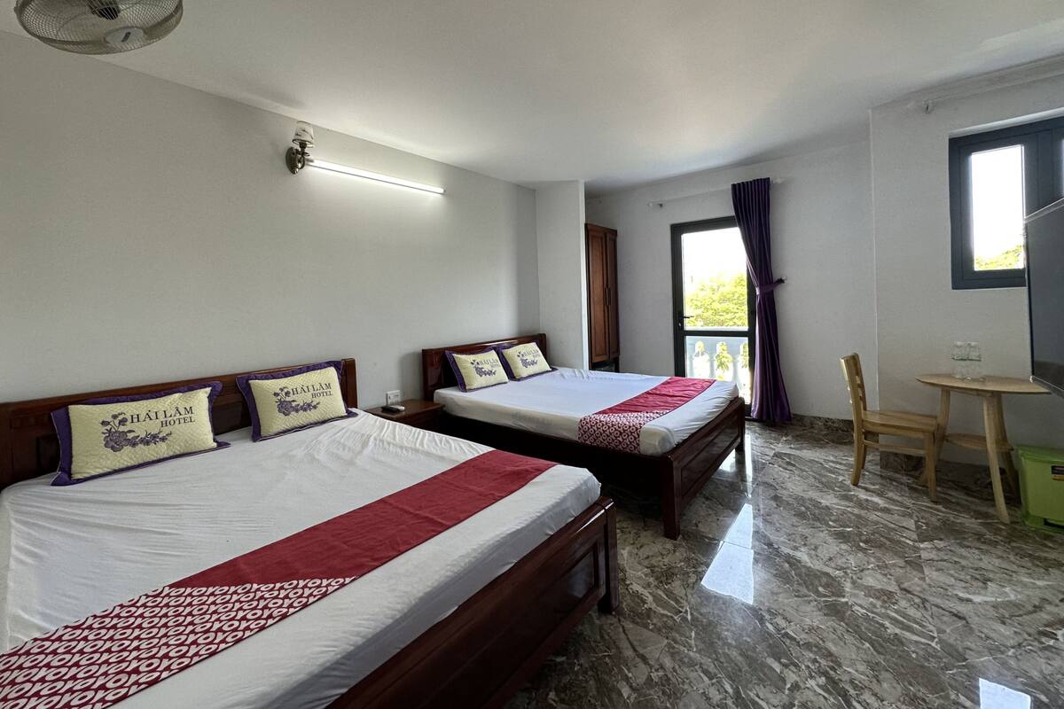 Ein Hotelzimmer mit zwei Betten, einem kleinen Tisch, einem Stuhl, einem Deckenventilator und einem Fenster mit Vorhängen.