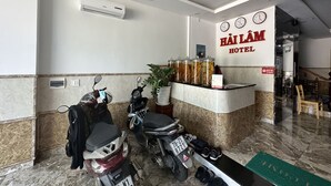 Een hotel receptie met een marmeren vloer, twee geparkeerde motorfietsen, een receptiebalie met een plant en een klok aan de muur.