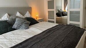 Un letto rifatto con cura, con una testiera grigia, cuscini bianchi e grigi e una coperta grigio scuro. Una lampada da comodino con un paralume giallo è posizionata sul tavolino accanto al letto.