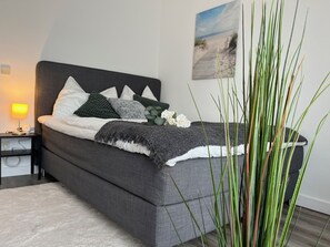 Een net opgemaakt bed met een grijze hoofdbord, witte en groene kussens en een grijze deken. Er staat een nachtkastje met een lamp en een kleine plant. Een grote groene plant staat op de voorgrond.