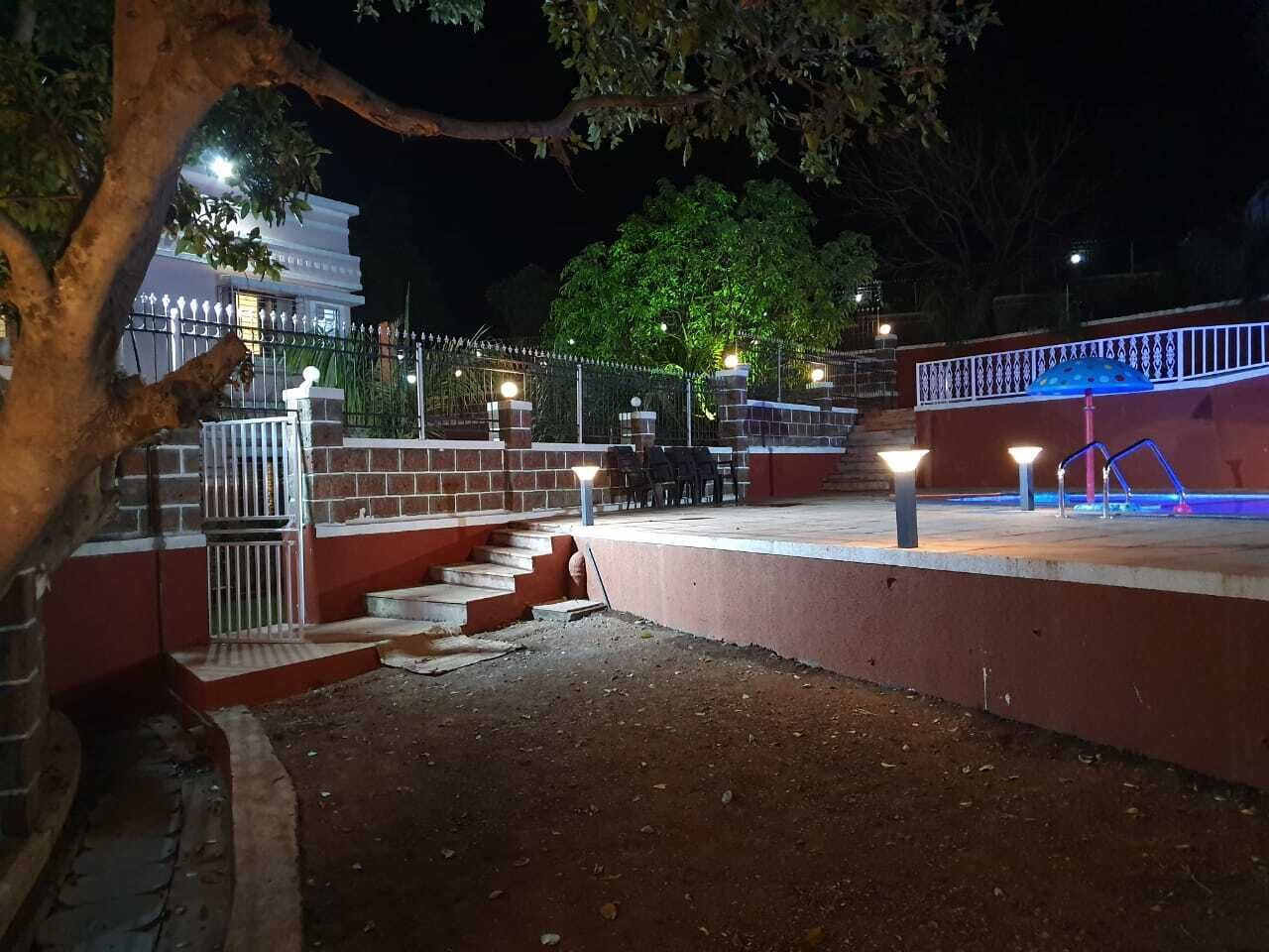 Un espacio exterior nocturno con escalones que conducen a una entrada con reja, una zona de piscina con un elemento acuático azul, y un edificio con señalización iluminada al fondo.