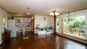 A spacious living area with a wooden floor, a kitchen with white cabinets, a dining table, and a large sliding glass door leading to a deck. - Badasol Fragrance Pension (Taean)