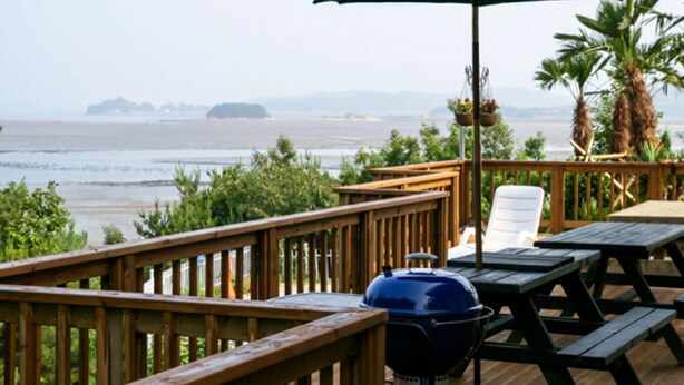 A wooden deck with picnic tables and a grill, overlooking a beach and ocean. - Badasol Fragrance Pension (Taean)