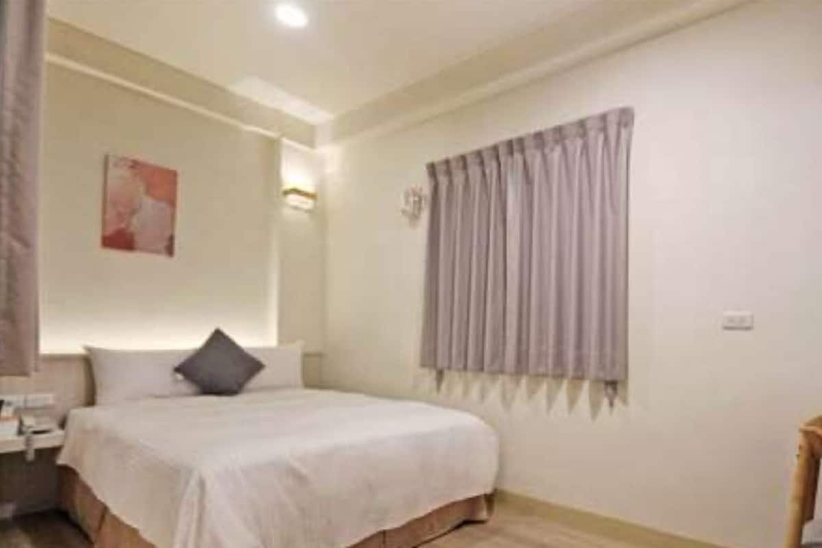 Un dormitorio con cama, mesita de noche, silla, ventana con cortinas y un cuadro en la pared.