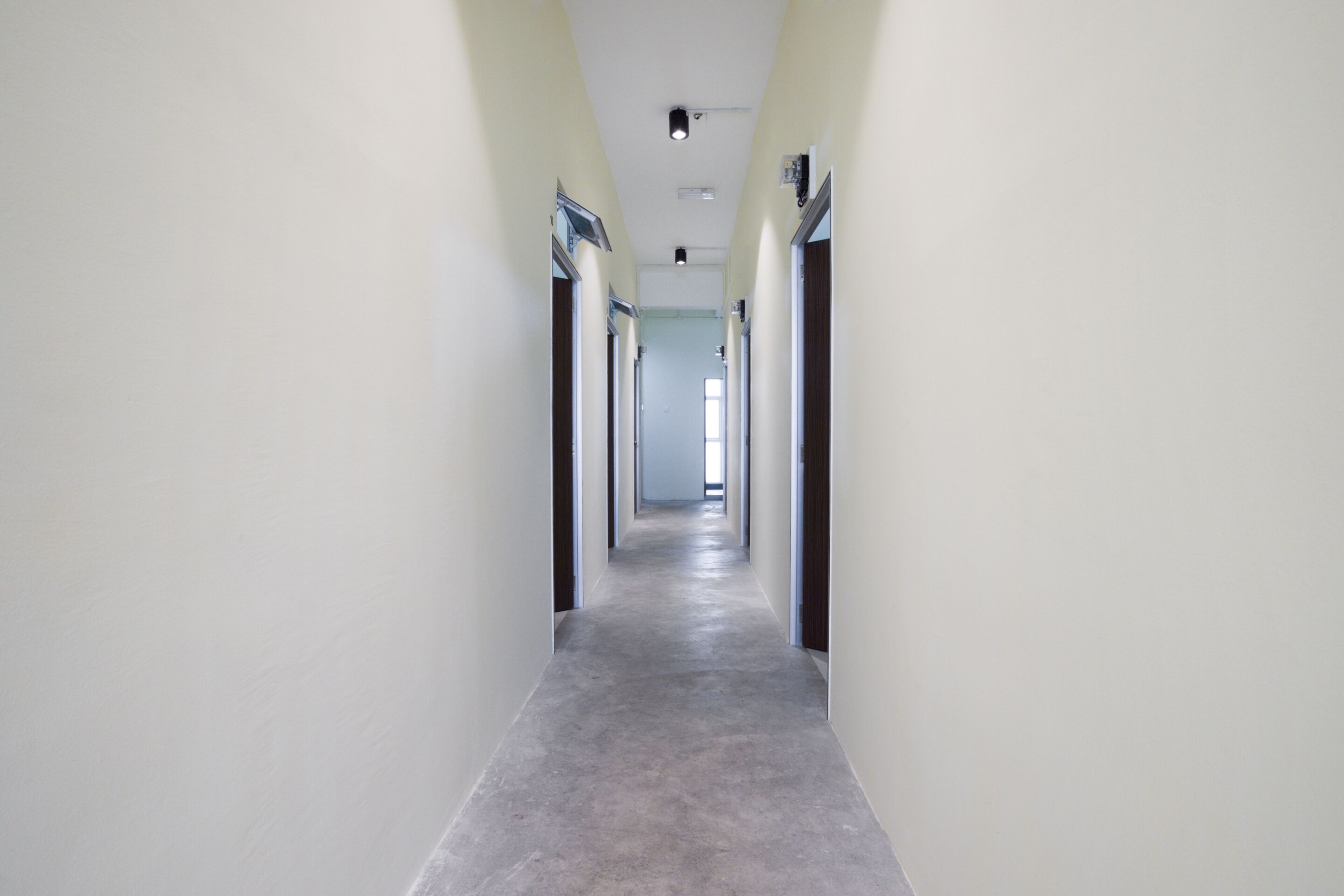 A long, narrow hallway with light-colored walls and a grey floor, leading to a door at the end.