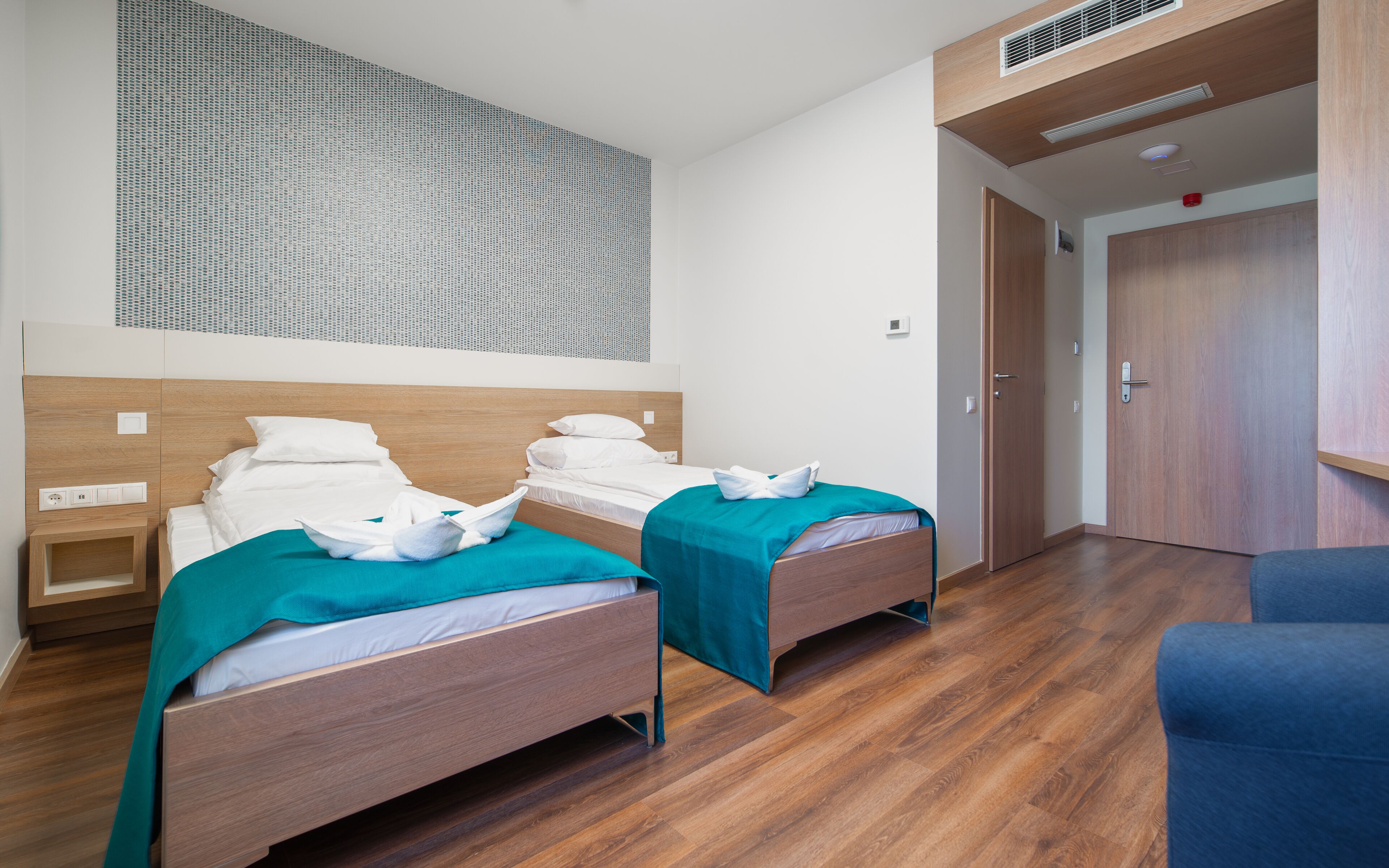 Een hotelkamer met twee eenpersoonsbedden, houten vloeren, een groot raam met een gedetailleerd uitzicht, en een deur naar een andere kamer.