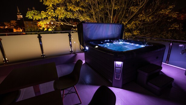 A hot tub with a control panel and a chair, set on a balcony with a railing and a view of trees and buildings. - Hotel Szeghalom (Szeghalom)