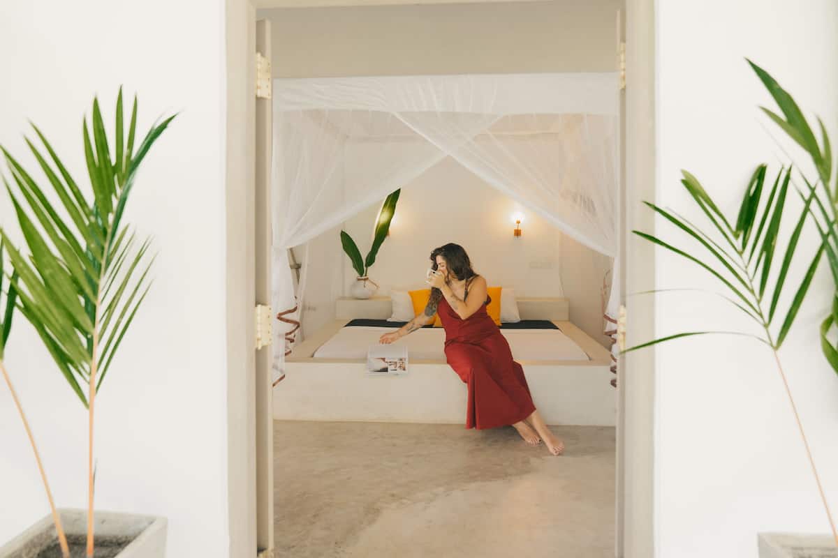 Une femme vêtue d’une robe rouge est assise sur un lit à baldaquin blanc, entourée de plantes en pot.