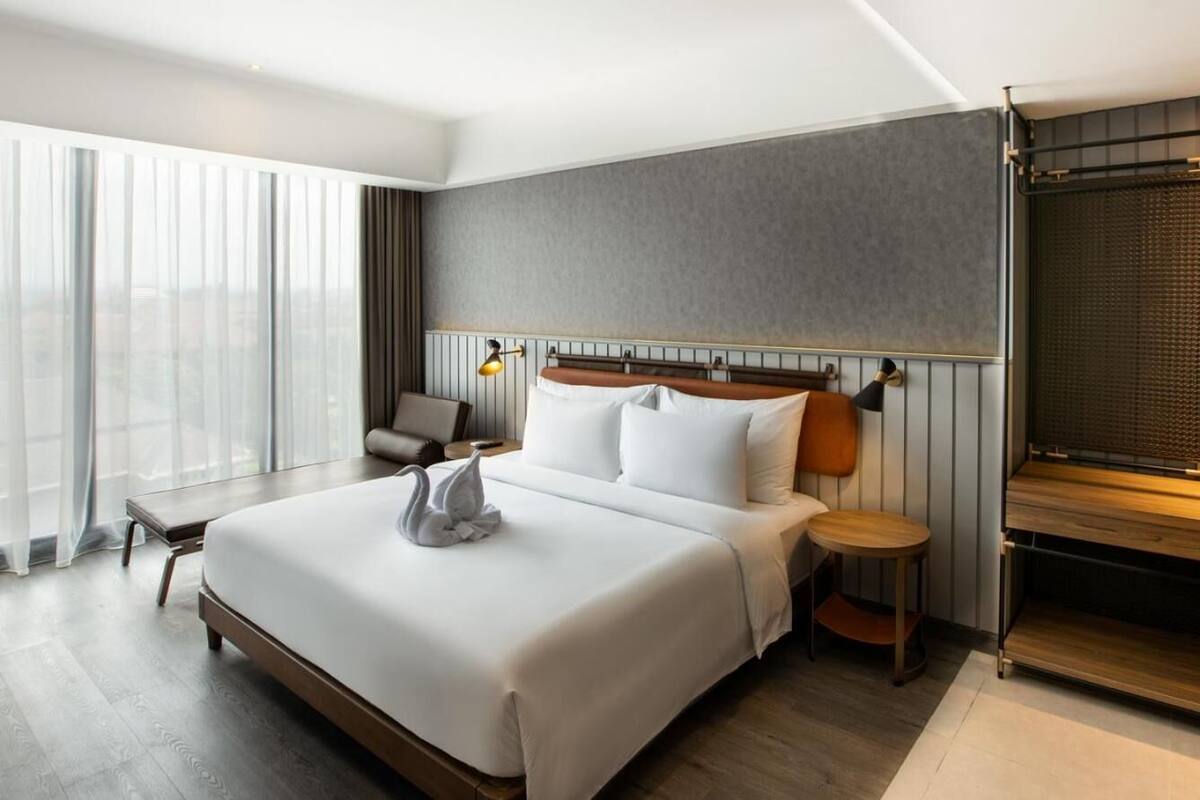 A modern hotel room with a large bed, a bedside table, a small round table, and a large window with sheer curtains.