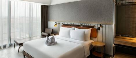 Une chambre d’hôtel moderne avec un grand lit, une table de chevet, une petite table ronde et une grande fenêtre avec des rideaux légers.