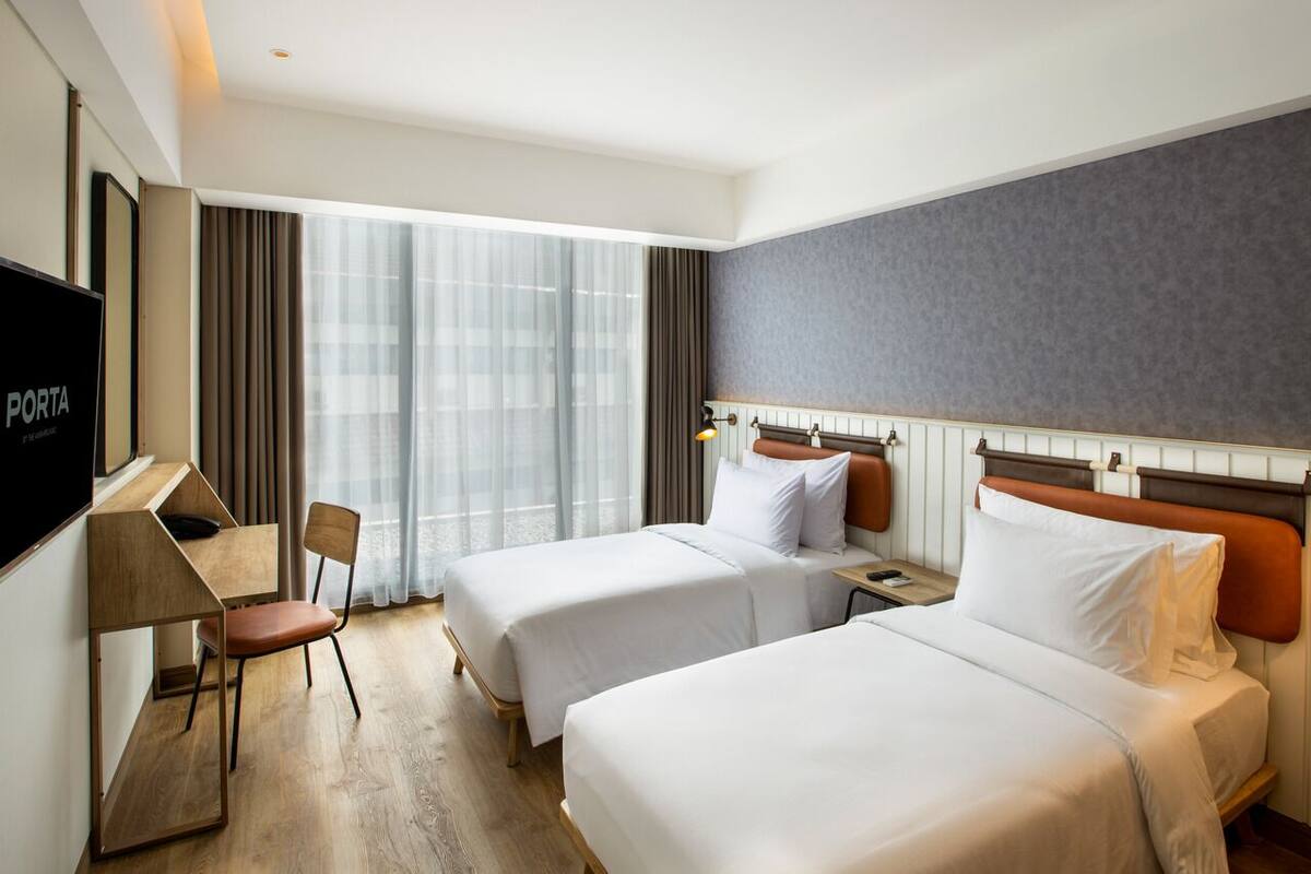 A hotel room with two beds, a desk, a chair, a TV, and a large window with curtains.