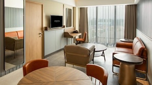 Habitación de hotel moderna con zona de comedor, sofá, televisor y ventana grande con cortinas.