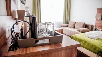 A wooden desk with a charging station, a bottle, and glass jars. A bed with a green blanket and a sofa are in the background.