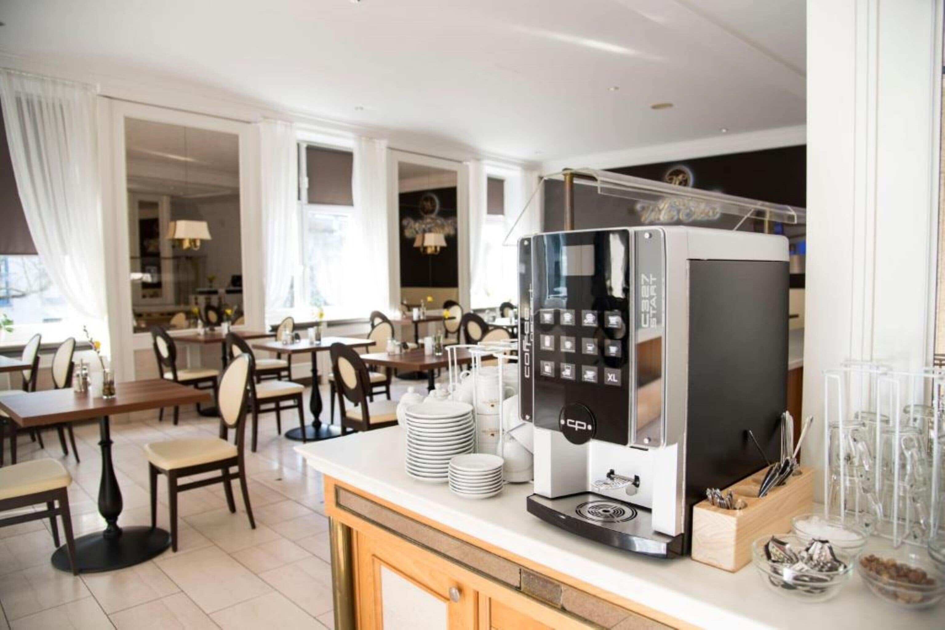 A modern hotel dining area with a coffee machine, tables, chairs, and a view of the kitchen.