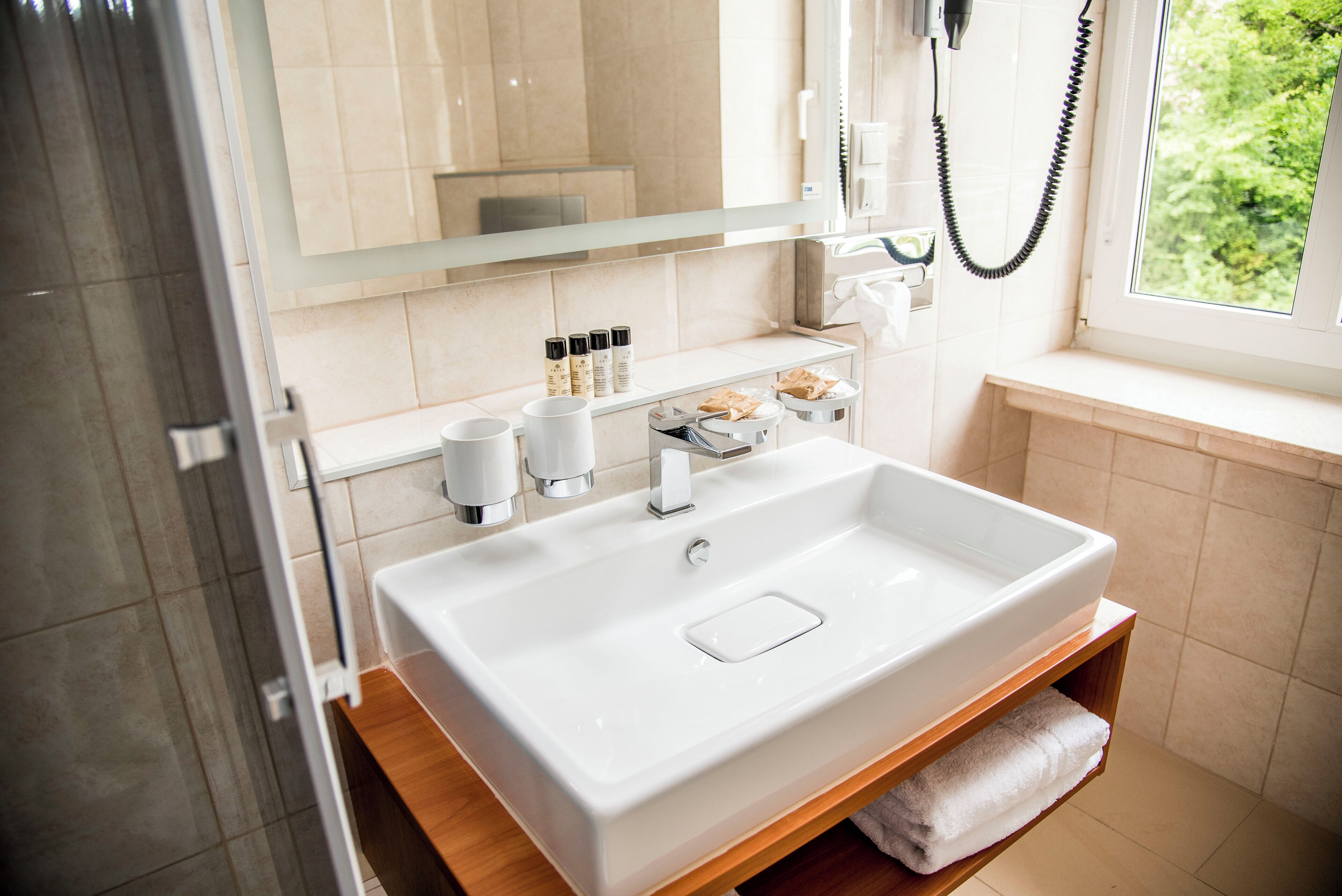 Une salle de bain moderne avec un grand lavabo blanc, un miroir et une fenêtre donnant sur un paysage verdoyant.