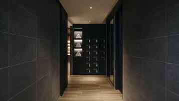 Un pasillo moderno con paredes de baldosas oscuras y piso de madera, que conduce a una habitación con una puerta oscura y estantes iluminados que exhiben objetos.