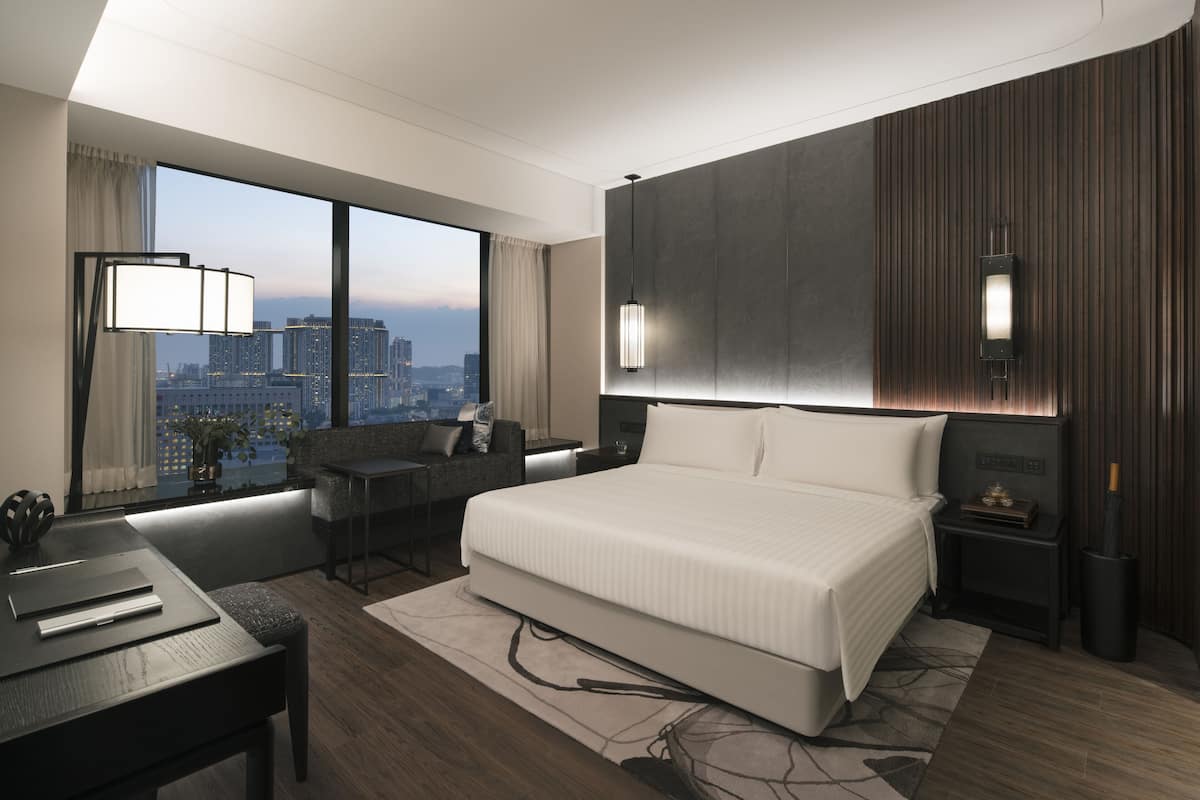 A modern hotel room with a large bed, a desk, and a view of the city skyline.