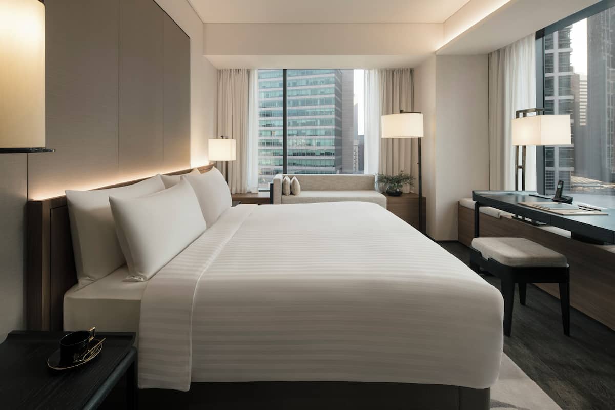 A modern hotel room with a large bed, a desk, and a view of a cityscape.