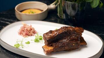 Un plato con un trozo de carne, un recipiente pequeño de salsa y algunas verduras sobre una superficie oscura.