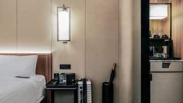 Habitación de hotel con cama, mesita de noche con lámpara, escritorio pequeño con computadora, maleta y minibar.