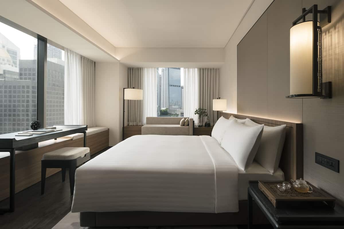A modern hotel room with a large bed, a desk, a chair, and a bedside table with a lamp.