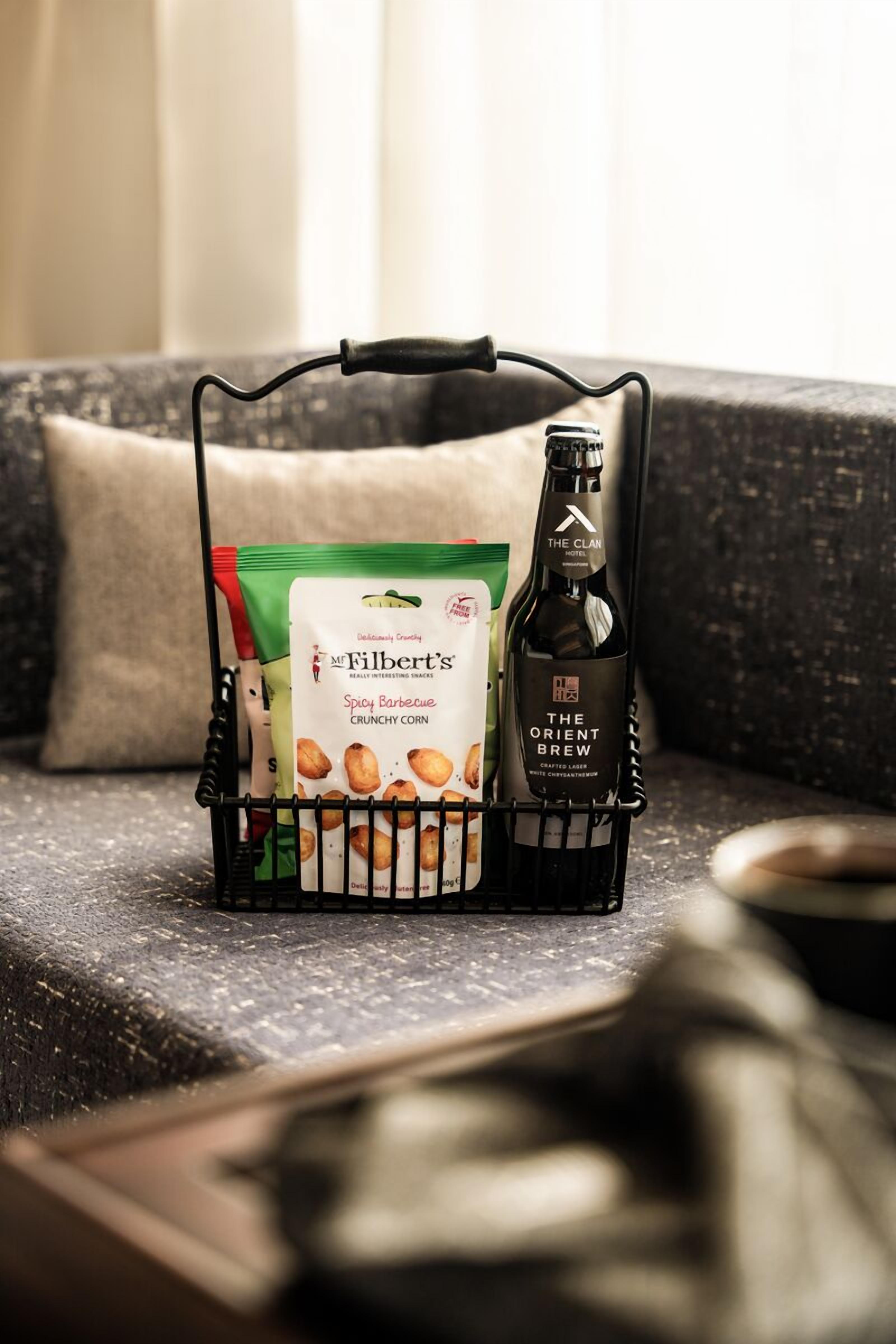 Una cesta negra sobre un sofá texturizado, con una botella de Orient Brew del The Clan Hotel y una bolsa de snacks Filbert's Spicy Barbecue Crunchy Corn.