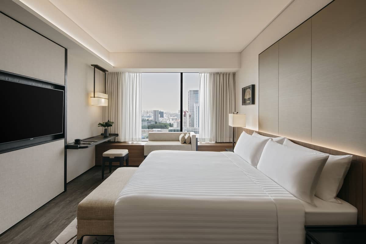 A modern hotel room with a large bed, a desk, and a view of the city skyline through floor-to-ceiling windows.