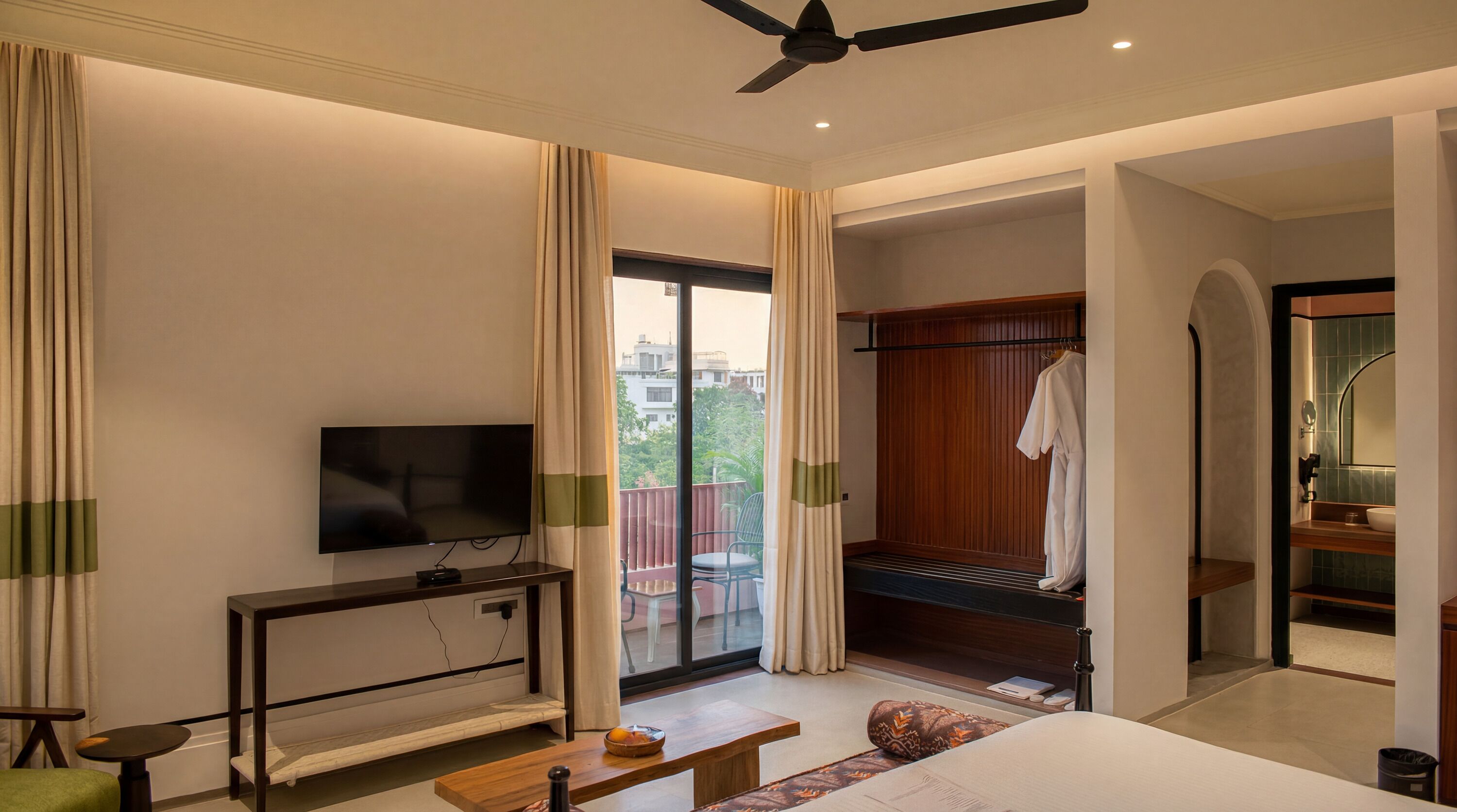 A modern hotel room with a flat-screen TV, a wooden console table, a sofa, a small table with a bowl of fruit, and a sliding door leading to a balcony.