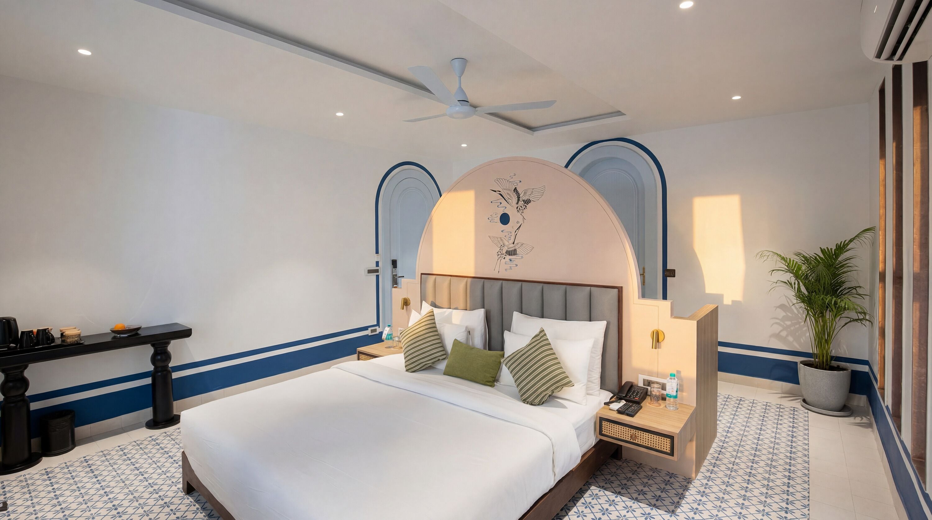 A hotel room with a bed, bedside table, lamp, and a potted plant.