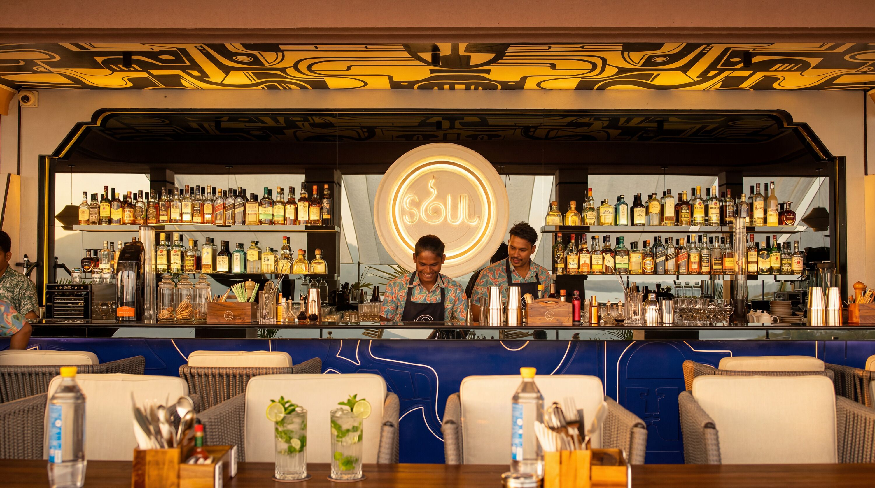 Zona de bar con un letrero de neón, estantes llenos de botellas de licor y taburetes.