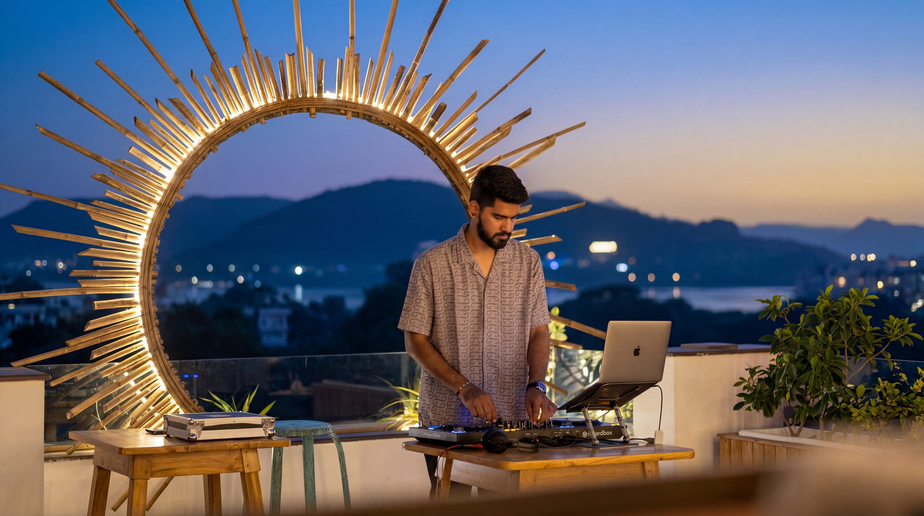 Una persona manejando un equipo de DJ con una computadora portátil en un balcón, con vista a un paisaje urbano y montañas.