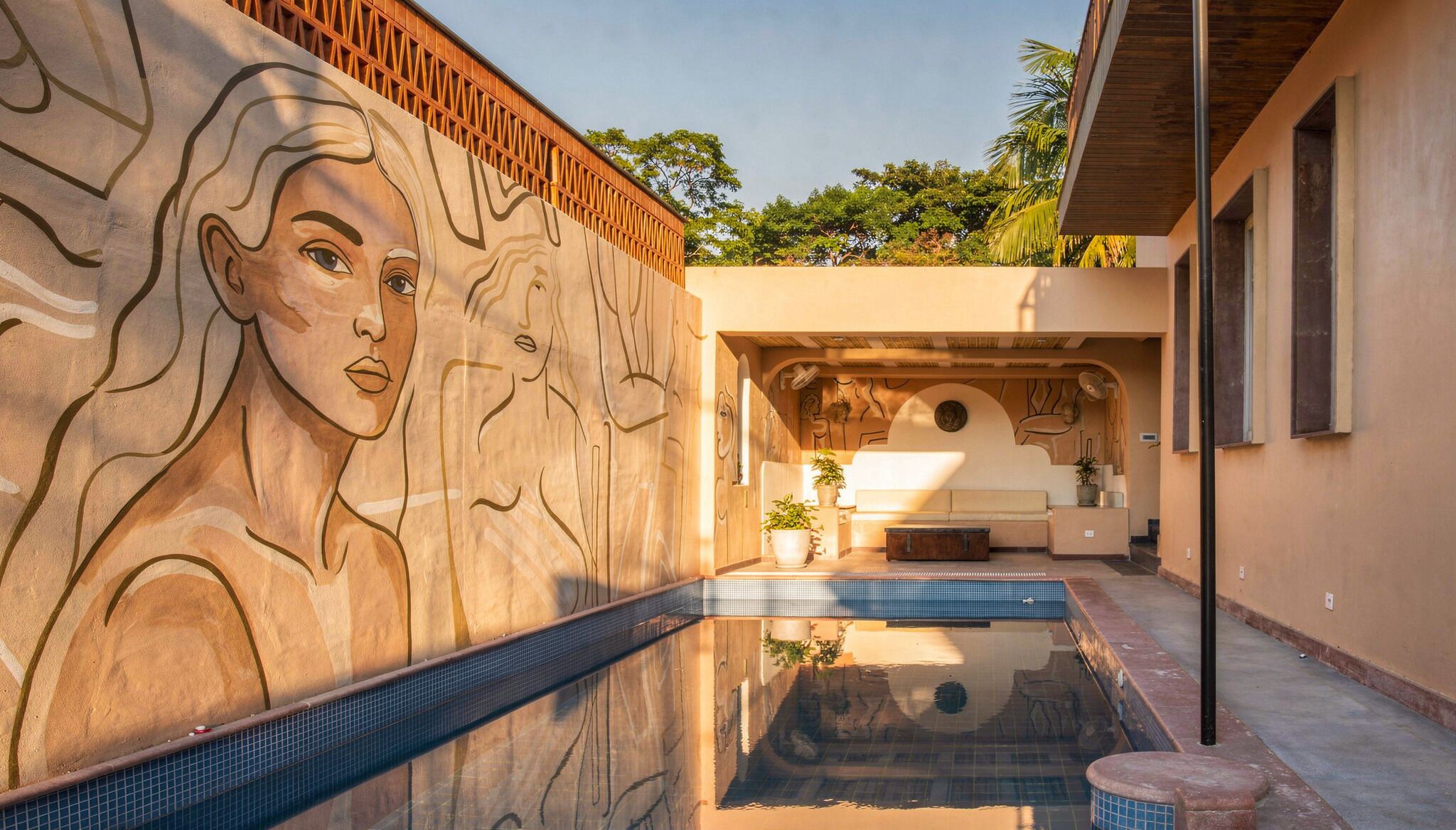 Una piscina con un mural, un área de descanso con un sofá y plantas, y una cerca de madera.