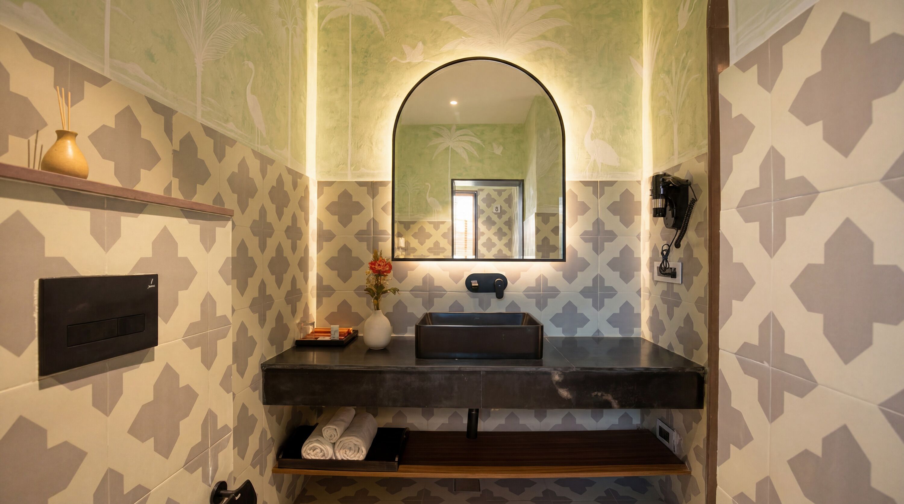 A bathroom with patterned walls, a mirror, a sink, a towel rack, and a shelf with a vase.