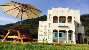 Un edificio blanco con detalles azules y ventanas arqueadas, una mesa de picnic y un paraguas grande.