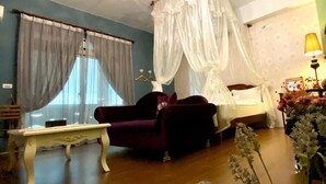Una habitación con cama bajo un dosel, un sillón morado, una mesita y una ventana con cortinas.