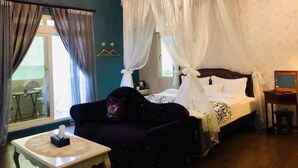 Un dormitorio con cama con dosel, un sofá oscuro, una mesita con una caja de pañuelos y una ventana con cortinas.
