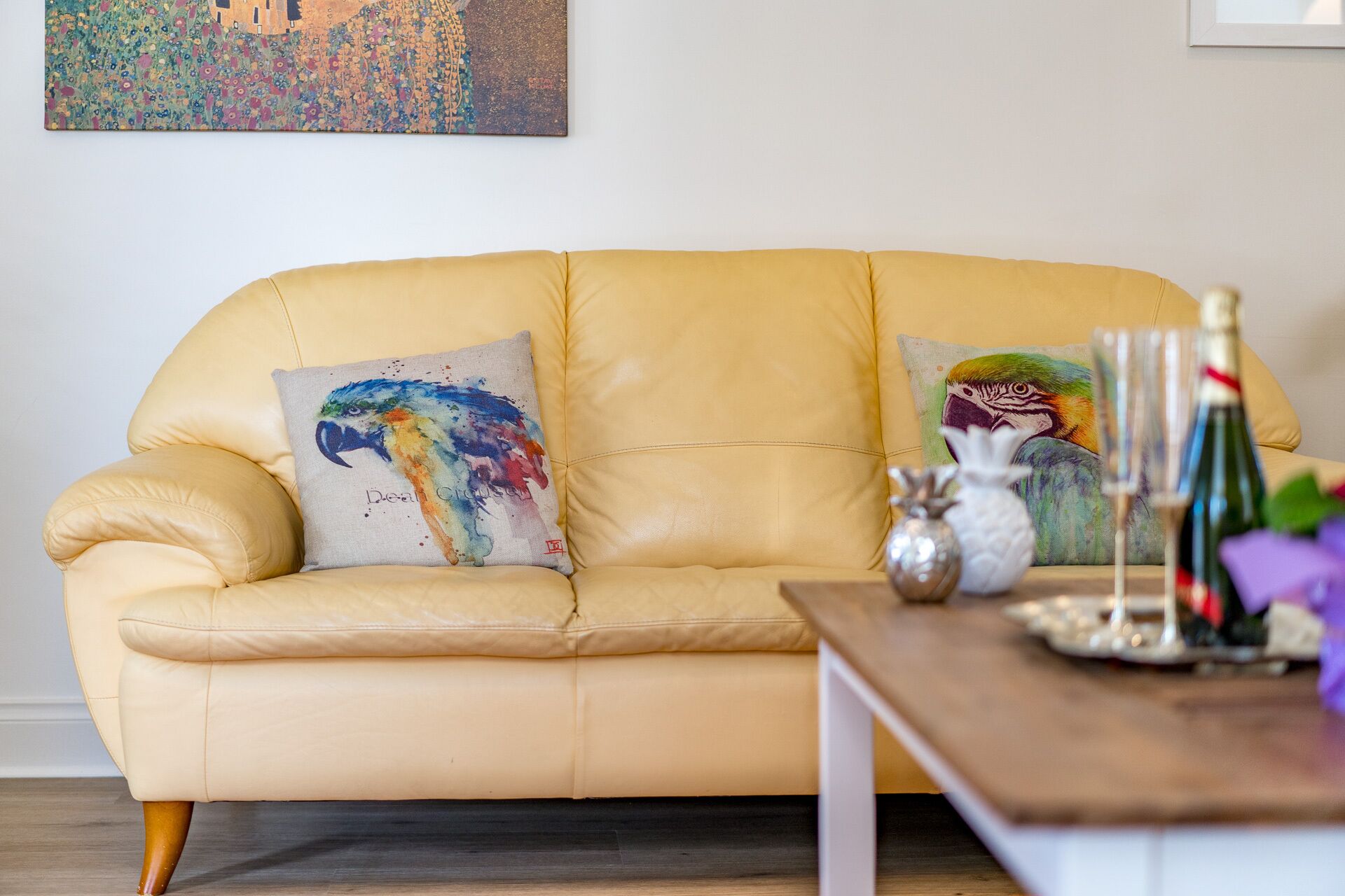A yellow leather sofa with two colorful pillows, a wooden side table with a bottle and glasses, and a painting on the wall.