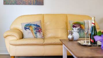 A yellow leather sofa with two colorful pillows, a wooden side table with a bottle and glasses, and a painting on the wall.