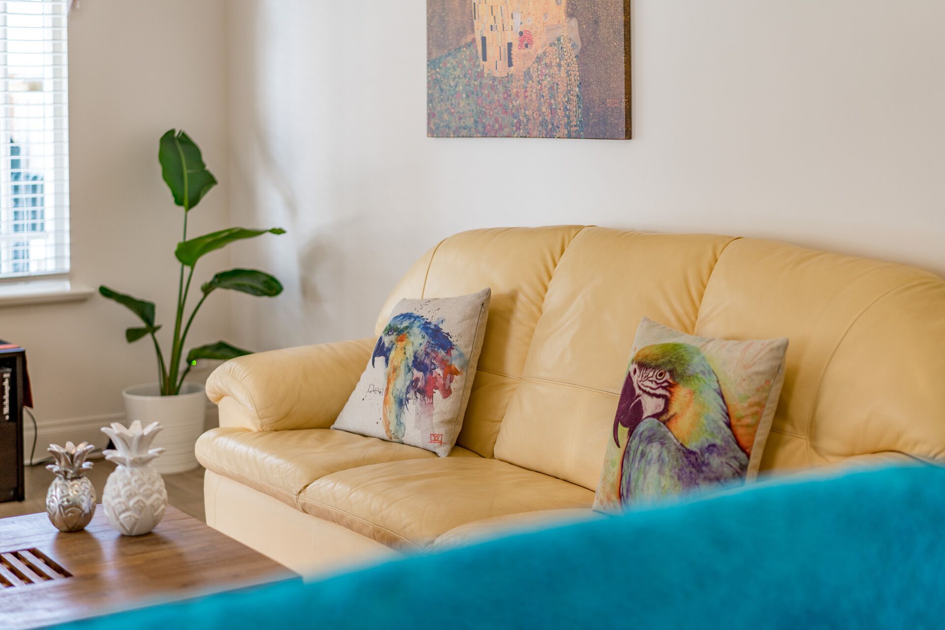 Un sofá de cuero amarillo con dos almohadas coloridas, una planta blanca en maceta y un cuadro en la pared.