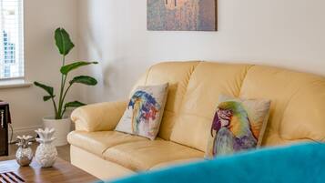 Un sofá de cuero amarillo con dos almohadas coloridas, una planta blanca en maceta y un cuadro en la pared.