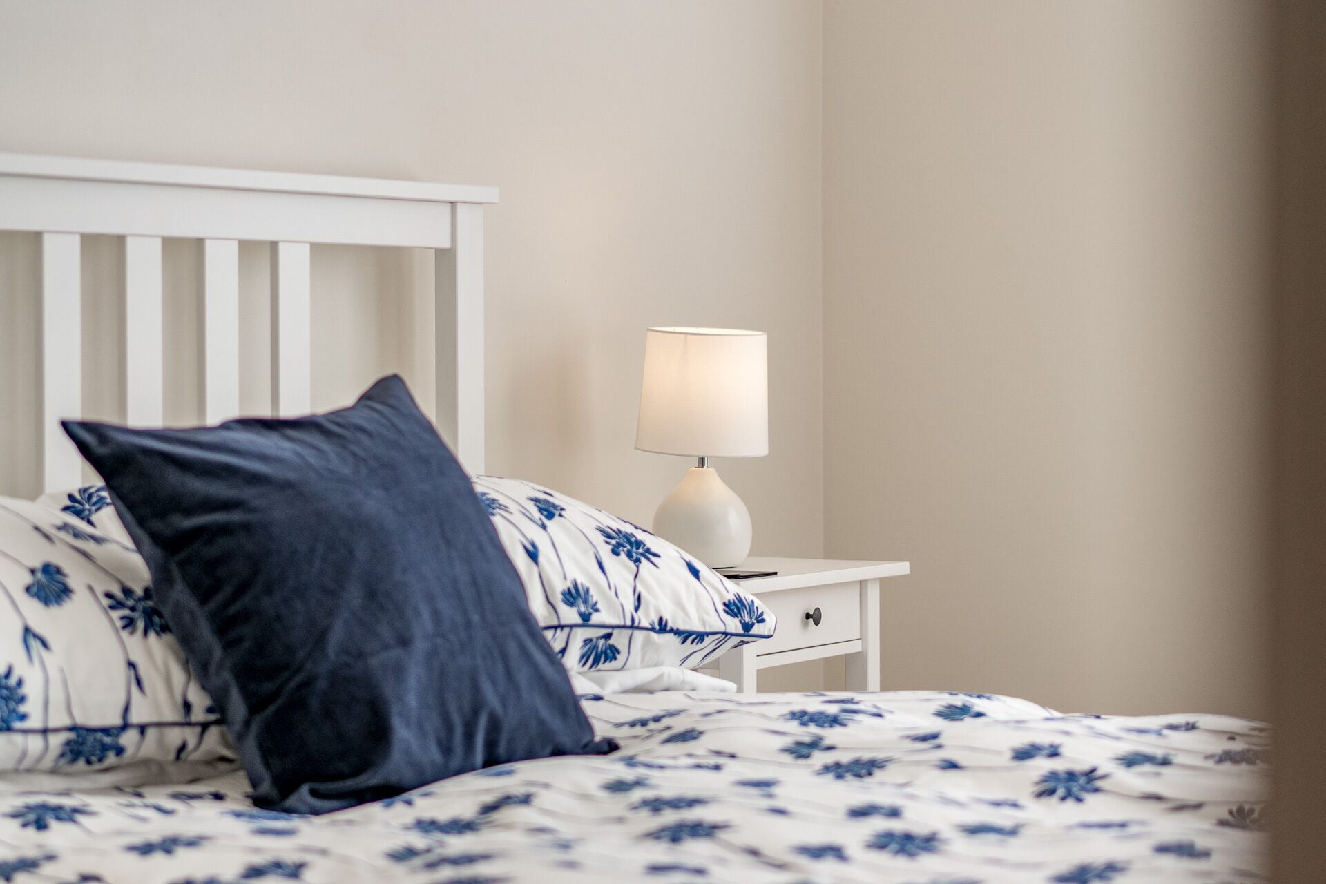 A neatly made bed with floral bedding, a white nightstand with a lamp, and a dark blue pillow.