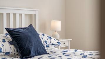 A neatly made bed with floral bedding, a white nightstand with a lamp, and a dark blue pillow.
