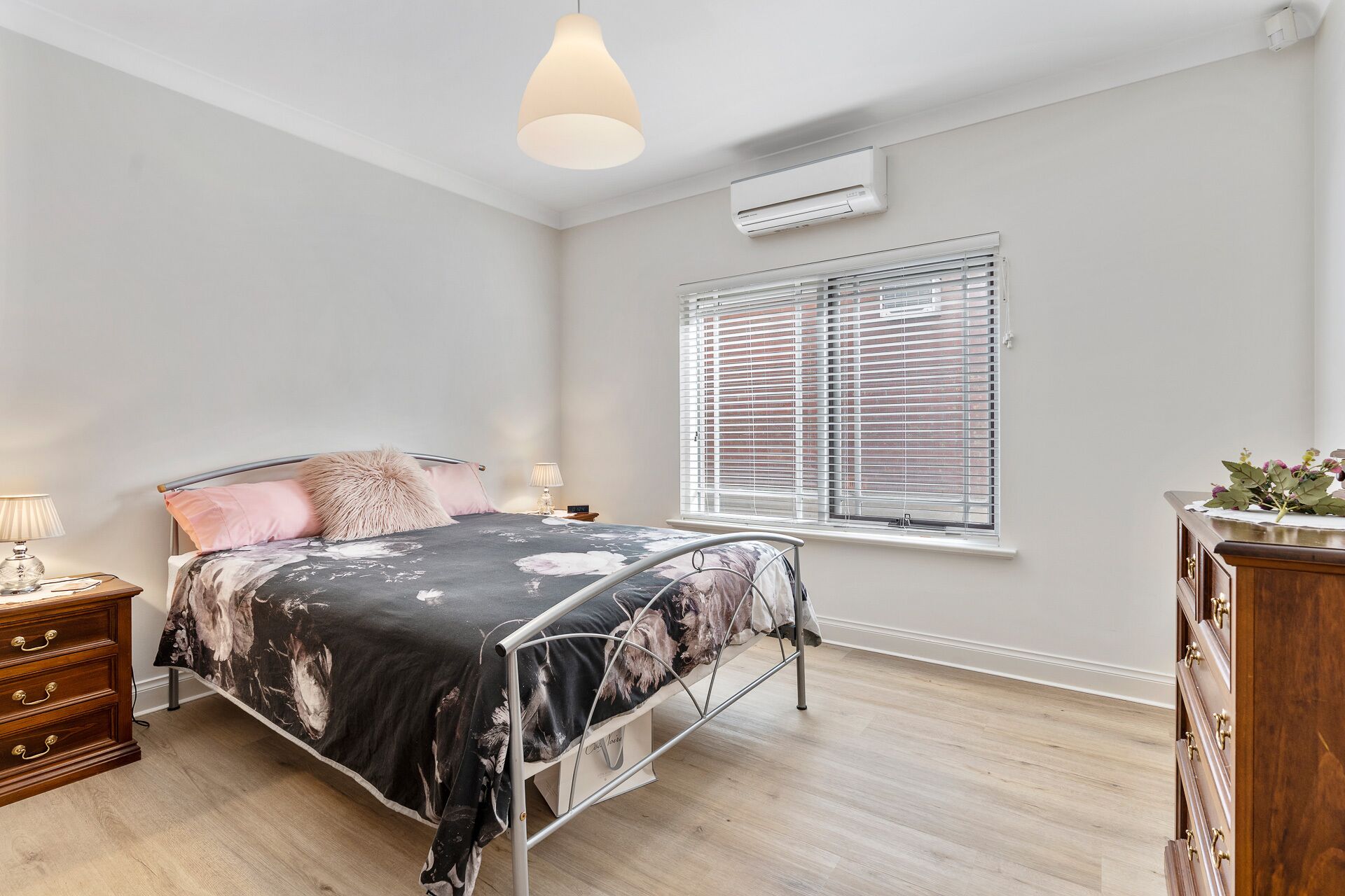 A bedroom with a metal bed frame, a wooden dresser, a bedside table with a lamp, and a window with blinds.